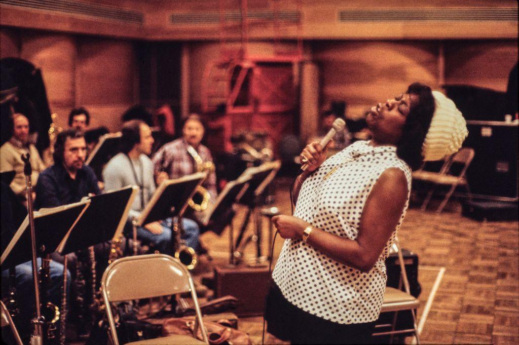 Sarah Vaughn in full flight, performing with a live orchestra. She's wearing a cream woolly hat with a polka dot sleeveless blouse.