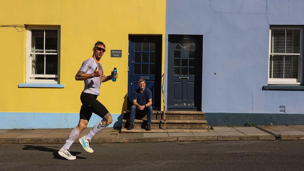 A competitor looks at the camera as he runs past a man dressed in blue shirt and jeans sits on the doorstep of a yellow house.
