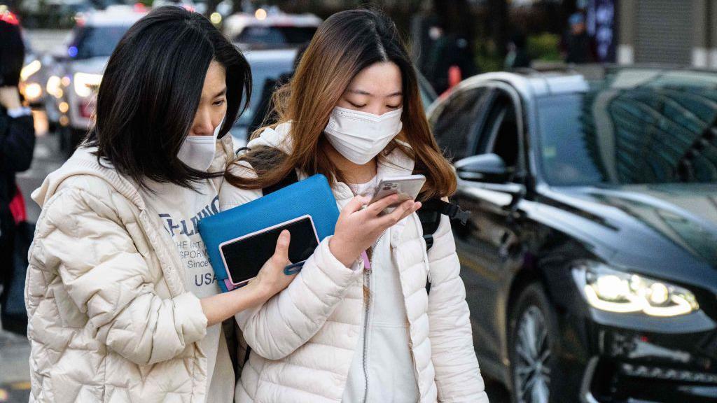 A student (R) arrives to sit for the annual college entrance exam, known locally as 'Suneung', outside the Ehwa Girls Foreign Language High School in Seoul on November 17, 2022. She is wearing a white puffer jacket, a mask and a backpack while looking at her phone. Next to her is an older woman, with her arm around her, holding a blue pouch and a phone.