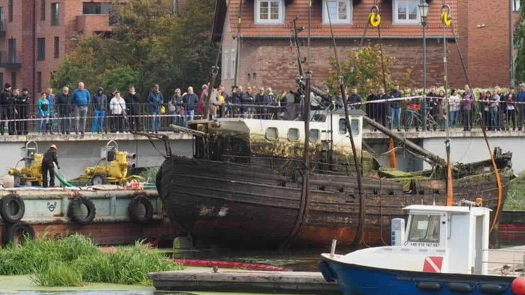 A is wooden fishing boat is being lifted out of the water by a large crane. above the boat behind a railing a number of people are looking on at the boat being lifted