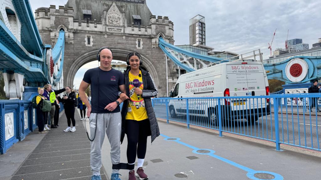 A man and woman linking arms and with a leg each tied together are walking across Tower Bridge. The woman has a pair of yellow Pudsey Bear ears on