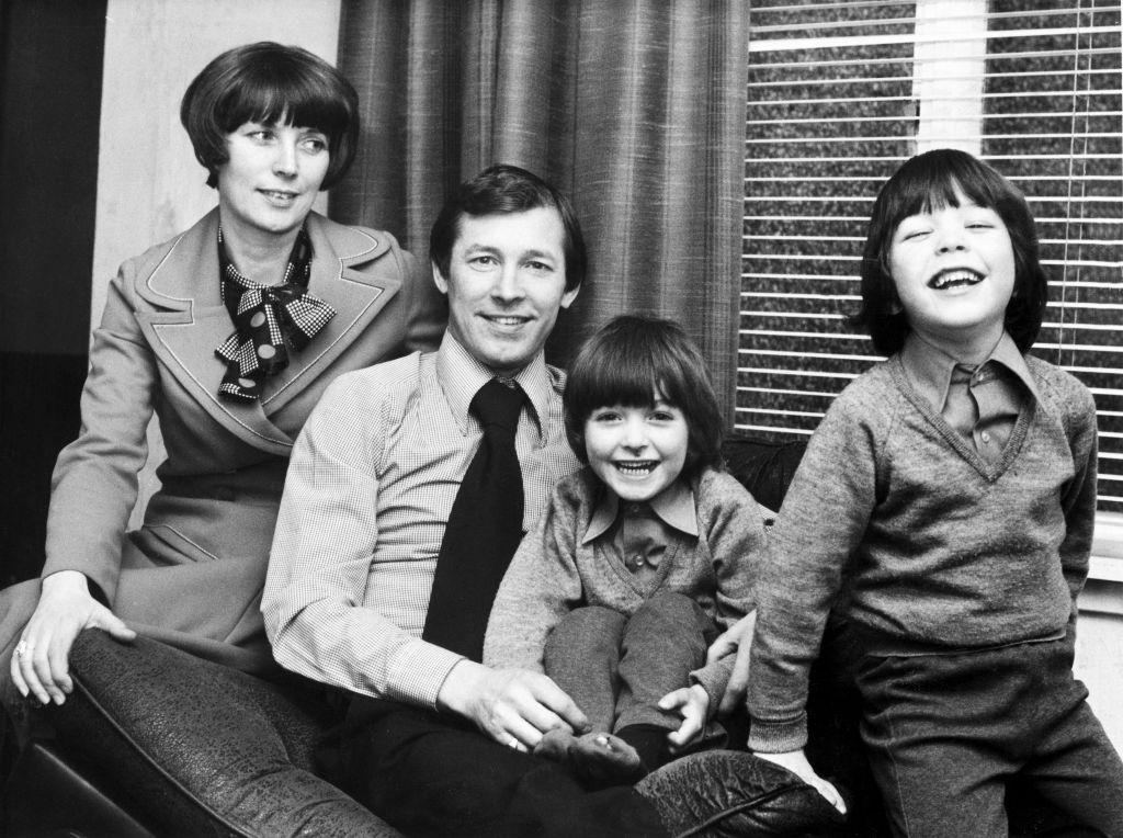 A family photo. Cathy Ferguson, Sir Alex and their two sons. The boys and Sir Alex look into the camera smiling, while Cathy looks at her family fondly.