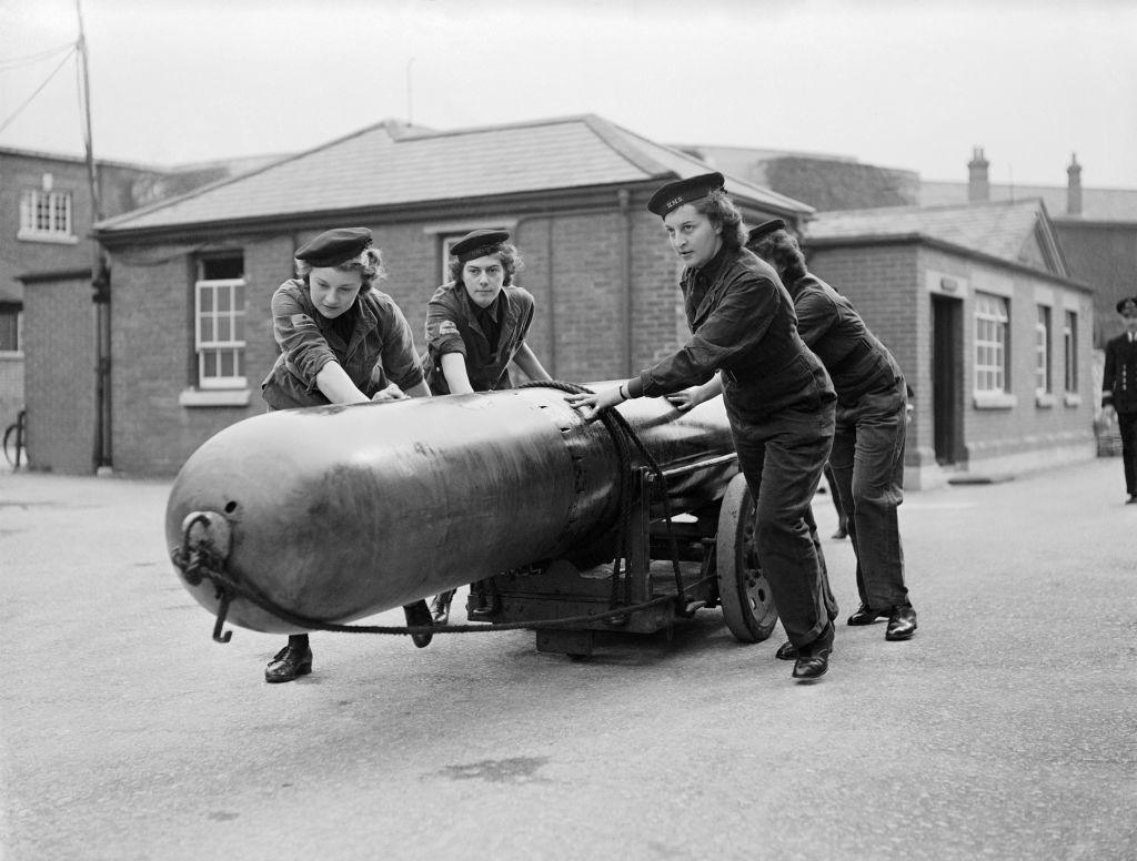 Four uniformed members of the Women's Royal Naval Service are seen pushing a torpedo across tarmac.