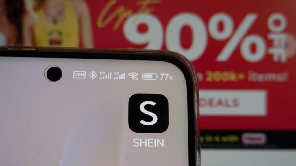 A smartphone screen displays the Shein app icon in the foreground, with a promotional advertisement offering discounts of up to 90% visible in the blurred background.