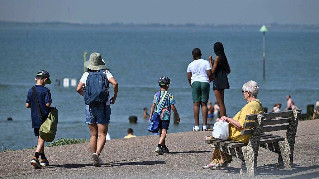 family walking along a coastal prominade.  one woman sitting on a bench looking out to sea.