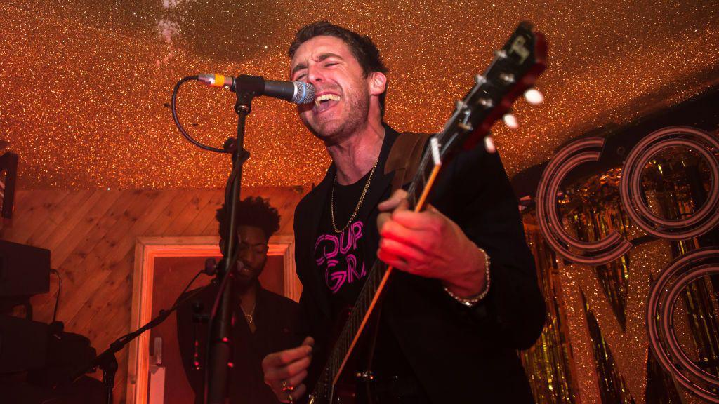 Miles Kane holds a guitar and sings into a microphone during a performance at the Moth Club