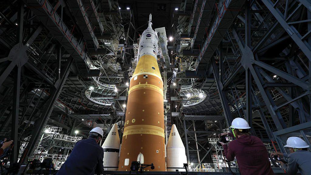 Artemis II sits in the Vehicle Assembly Building at NASA’s Kennedy Space Center as three workers wearing white hard hats look up at the rocket 