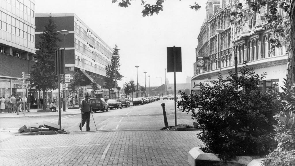 A black and white photo of The Hayes in Cardiff in 1983, with a road running through the middle, cars parked on it and driving through, and shoppers on pavements and crossing the road