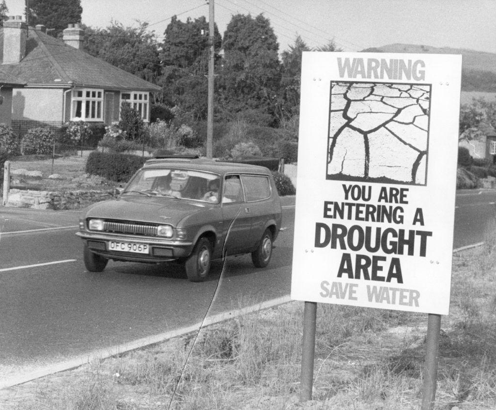 Black and white photo of a car on the road and a large sign saying 'warning, you are entering a drought area - save water'.