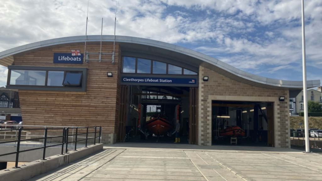A wood and stone building. It has a sign saying Cleethorpes lifeboat station. Inside are two orange lifeboats. there are blue skies with some cloud.