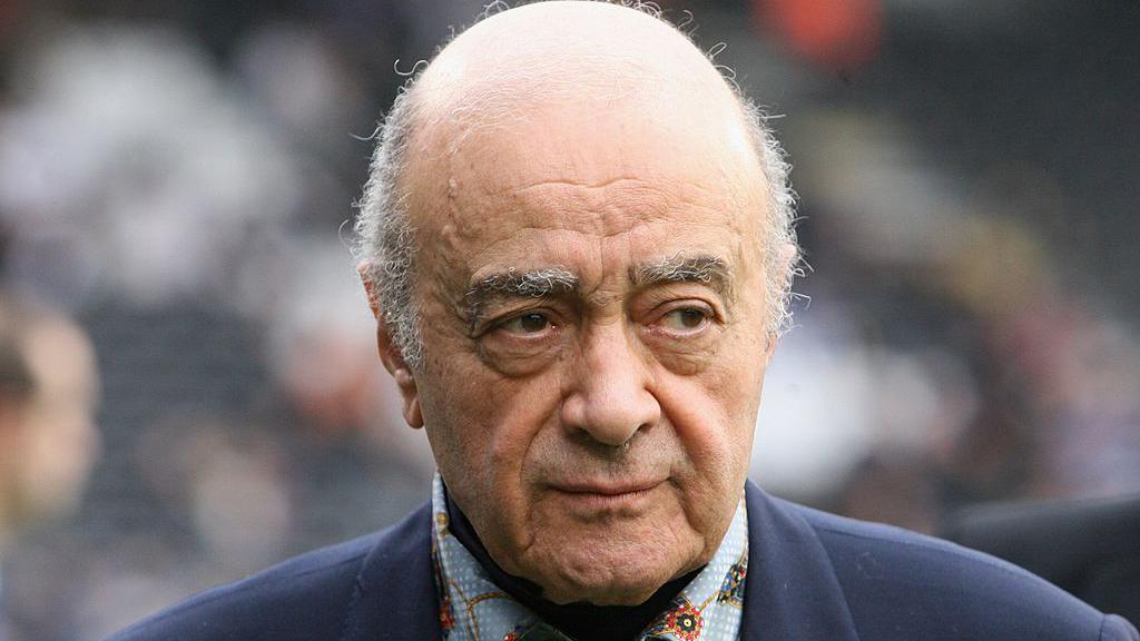 Mohamed Al Fayed, wearing a navy jacket and a colourful patterned shirt, pictured with a long lens at a Fulham FC match. He is facing the camera but his eyes are looking to his left