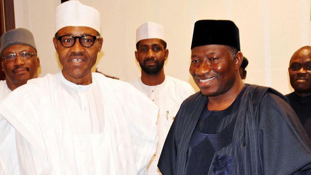 Muhammadu Buhari, in white traditional Nigerian clothes, and Goodluck Jonathan, in dark blue traditional clothes, stand next to each other smiling.
