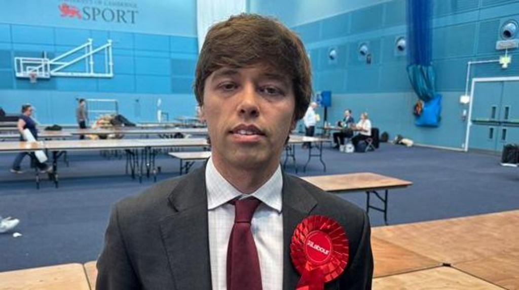 Cameron Holloway stands in a sports hall. He is wearing a suit and red rosette and has thick brown hair.