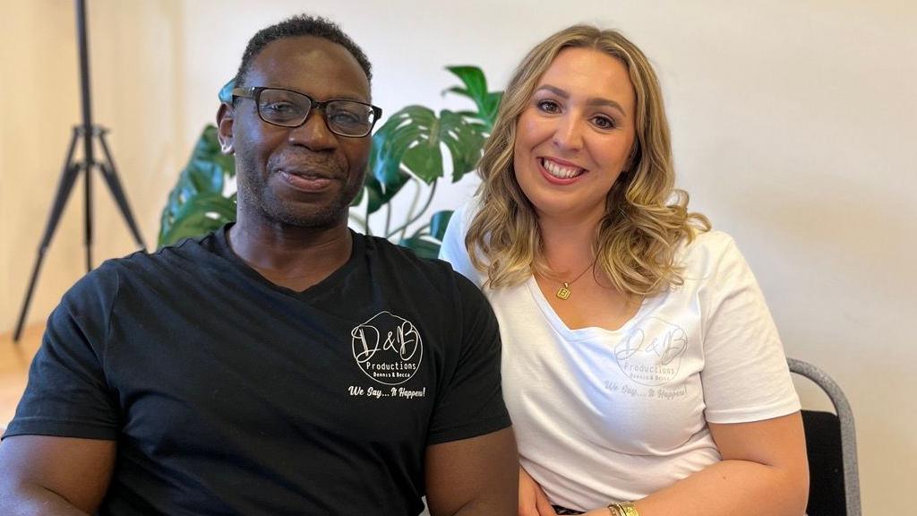 A man and woman are smiling and sitting next to each other with plants behind them.