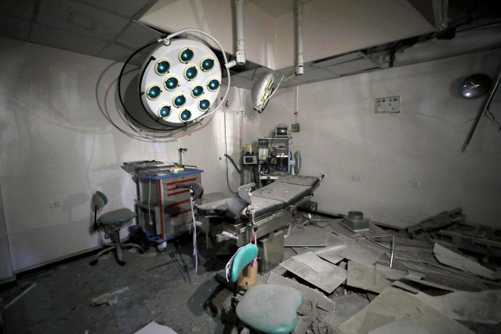 A room in the clinic contains a couch and chairs that are damaged and covered in a thick layer of dust. There is debris scattered on the floor.