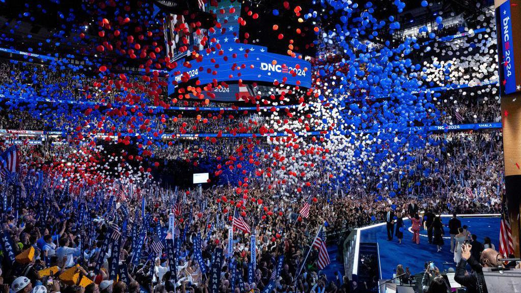 Balloons fall from the ceiling of an events centre in red, white and blue colours, with people cheering and celebrating