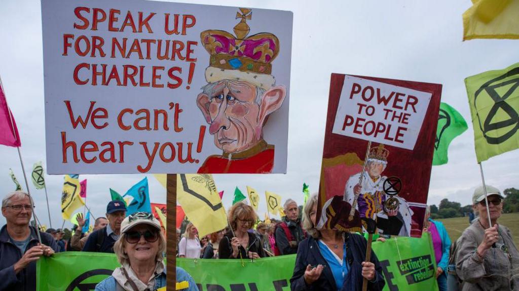 A crowd of campaigners hold placards and flags. A prominent sign reads "Speak up for nature Charles! We can't hear you!" alongside a cartoon of King Charles.