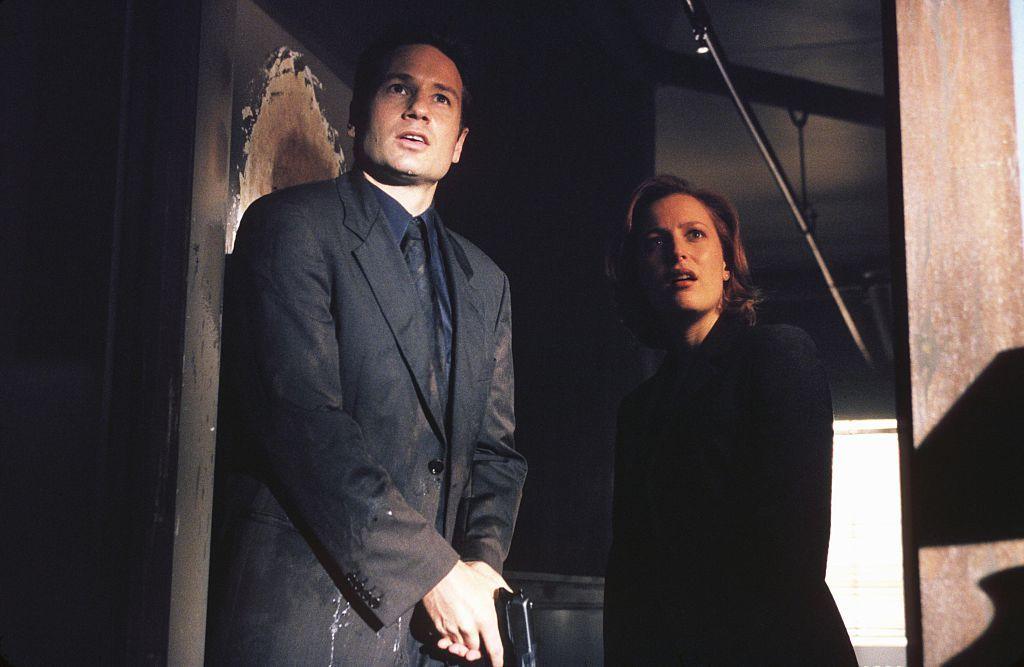 Mulder and Scully from The X-Files, in business suits, are standing in a doorway. Mulder, on the left, is carrying a handgun.