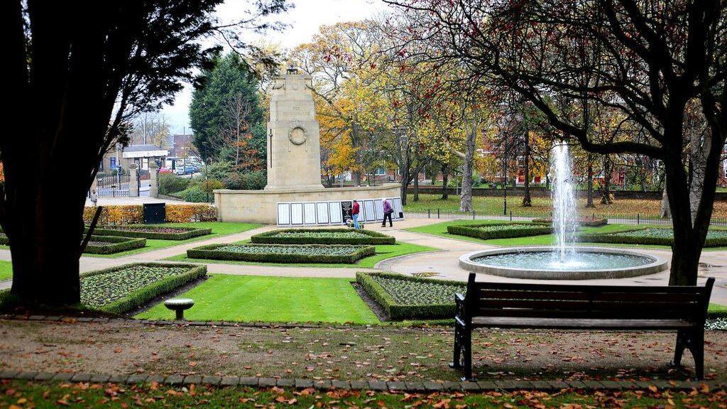 The cenotaph in Rotherham's Clifton Park, seen from across the memorial garden and fountain. Pedestrians are walking near the memorial and stopping to read. 