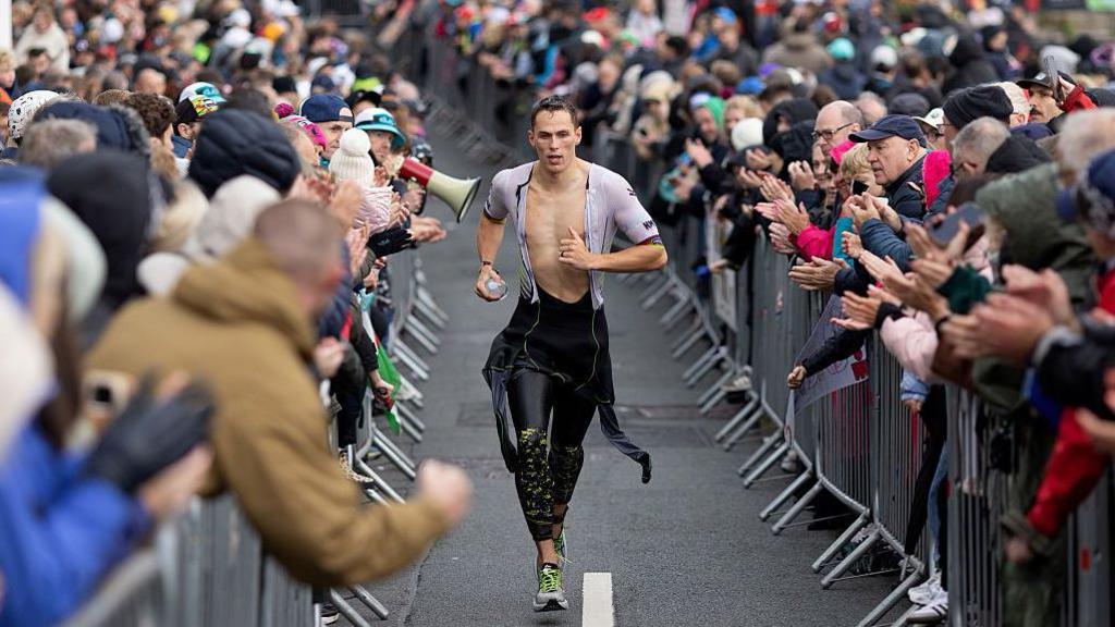 A man runs with his wetsuits unzipped to his waiste as he runs between crowds of people standing behind barriers