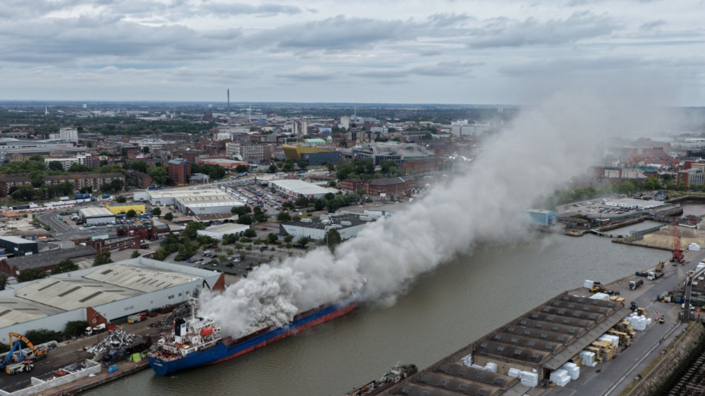 A blue and red ship in a dock which has lots of grey smoke billowing out from a fire. The Hull skyline is visible in the distance.