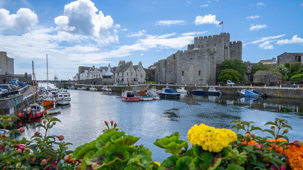 Castletown Harbour on a bright day. There are boats moored in the inner harbour and Castle Rushen can be seen on the right-hand side. There are colourful flowers in the foreground and blue sky with fluffy white clouds above.
