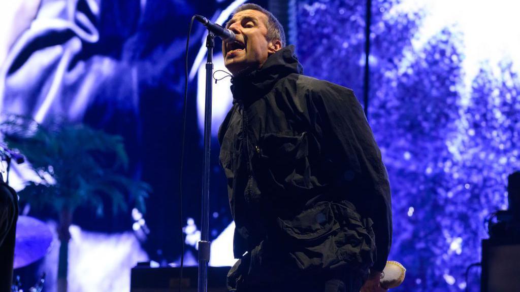 Liam Gallagher singing into a microphone, he is wearing a black jacket and has black hair. He is standing in front of a purple background.