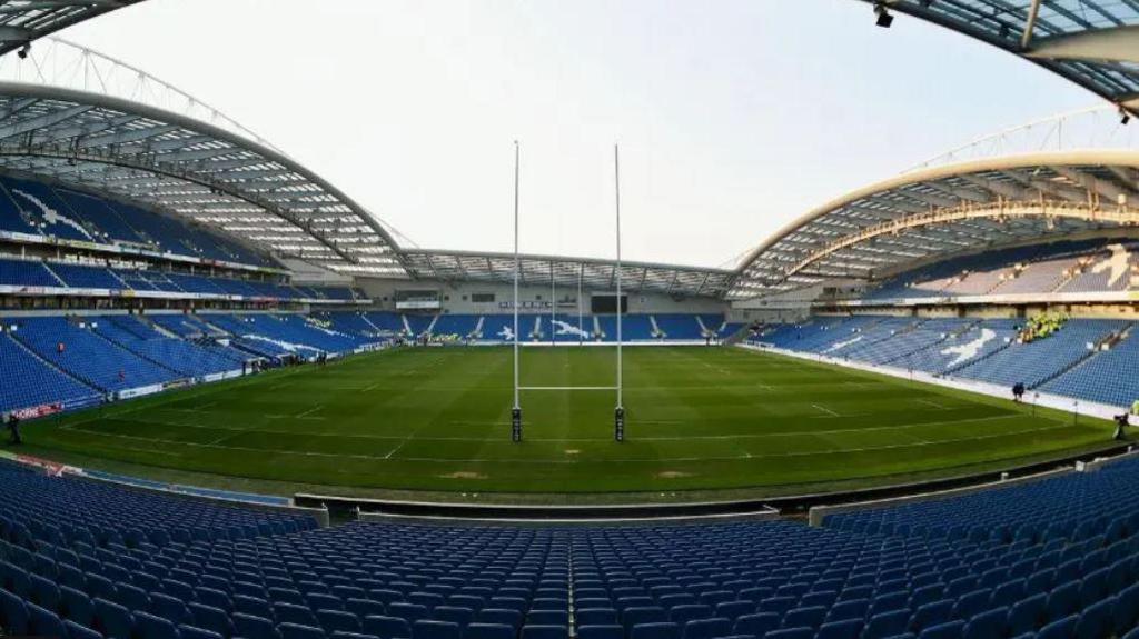 Wide shot of empty Amex Stadium in Falmer with rugby posts on the green pitch and thousands of empty blue seats around the edges.