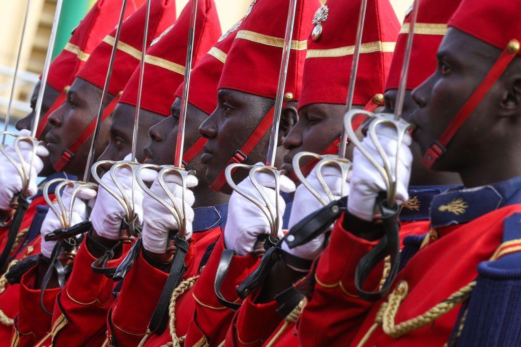 Soldiers dressed in red stand to attention and present their swords at a ceremony in Thiaroye Camp.