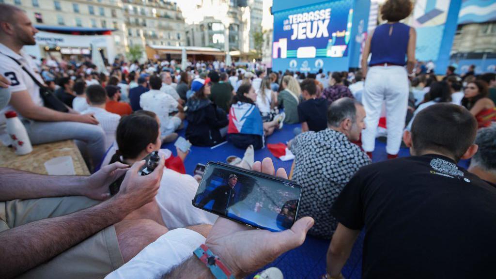 A hand in a crowd holds up a smartphone that shows Leon Marchand