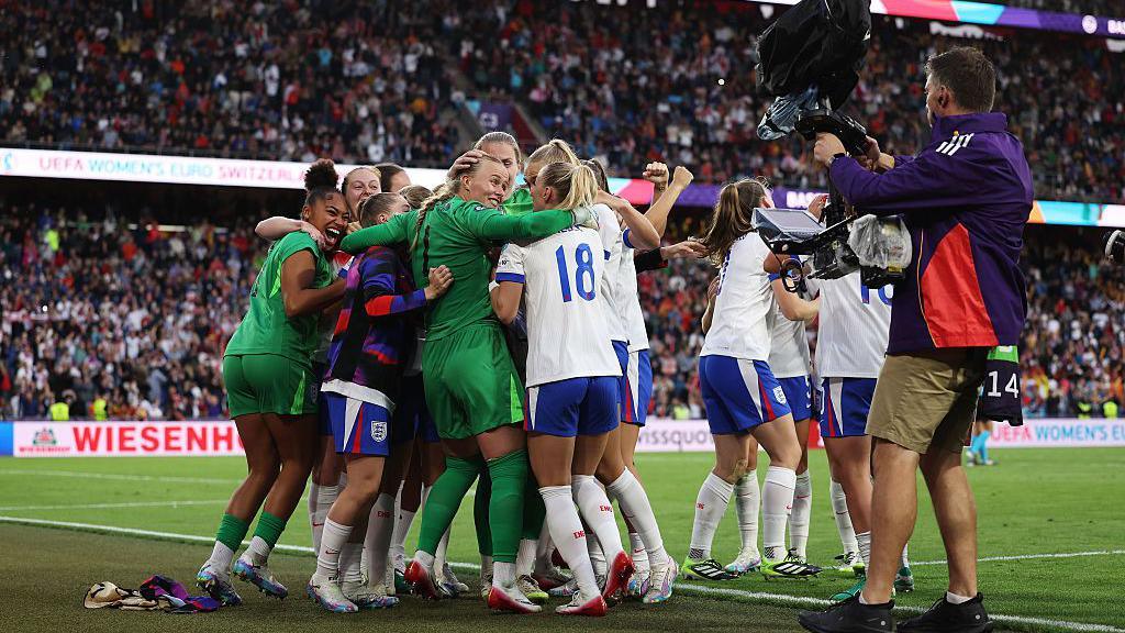 Hannah Hampton celebrates with her team after Chloe Kelly scores the team's winning penalty during the UEFA Women's EURO 2025 final on Sunday. The women are standing together in a group, hugging. Crowds are in the stands behind them and a photographer is taking a picture.