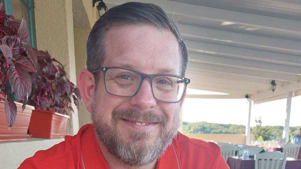 Andrew Rayner is in just off centre to the left looking towards the camera smiling. He has short dark grey hair and is wearing glasses and a red T-shirt. In the background are rows of tables and chairs.