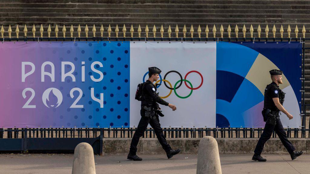 Two armed police officers walk in front of a poster for the Paris Olympics. They are seen in a side profile. 