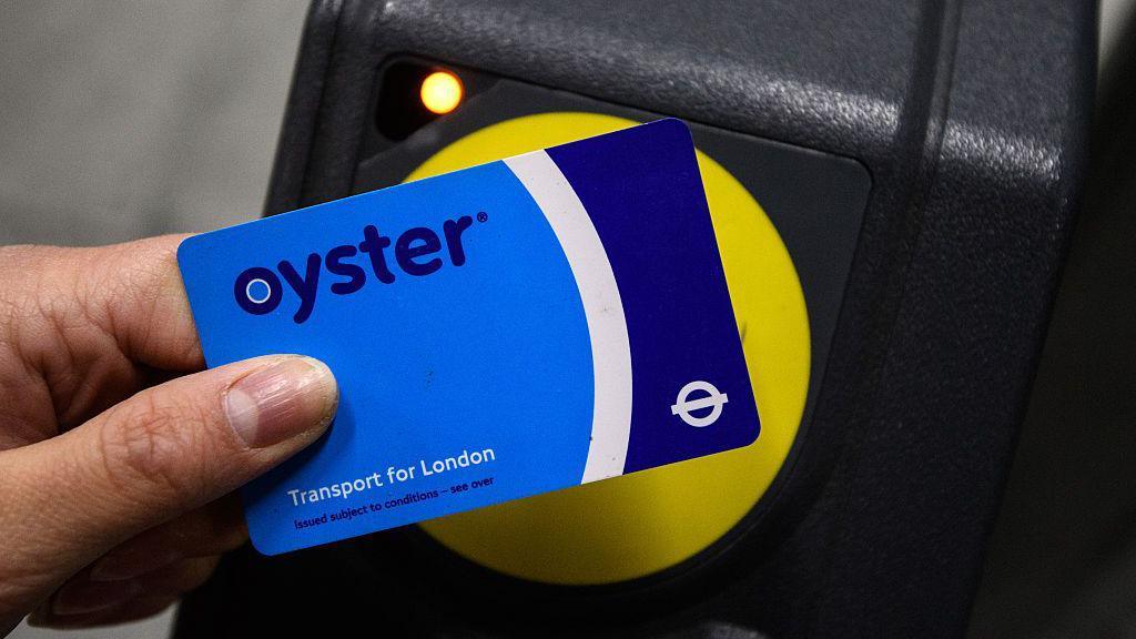 A close-up file image of a person tapping their blue and white Oyster card against the round yellow reader at a ticket barrier in a station building.