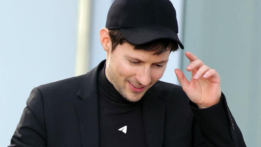 Pavel Durov, a white man white short black hair. He has designer stubble. He is wearing all black, including a cap, a turtleneck and a suit jacket.