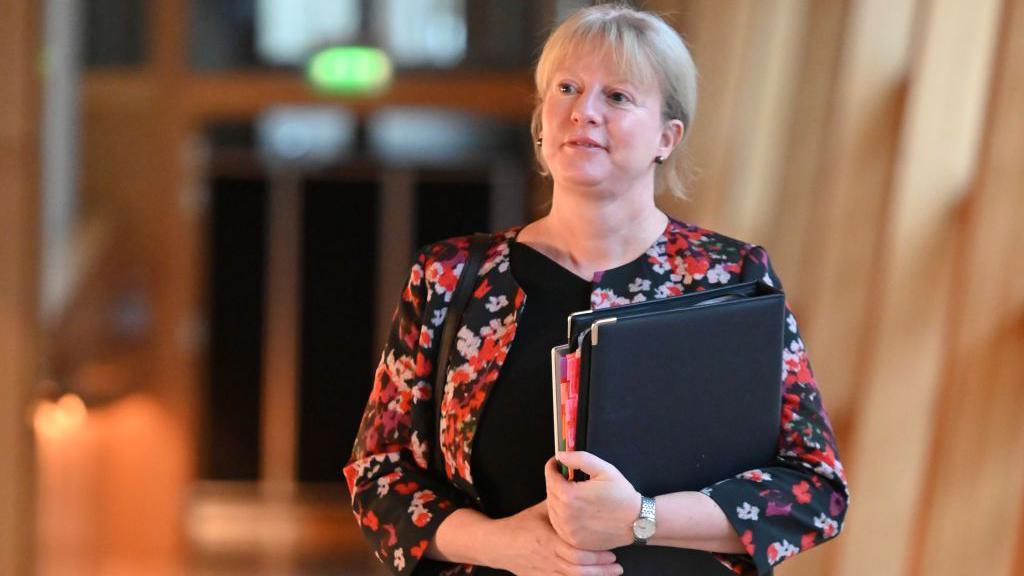 Shona Robison in a patterned red, white and black top, is carrying a black leather folder which is full of documents. She is smiling slightly and walking down a corridor in the Scottish Parliament. The background is blonde wood and glass.