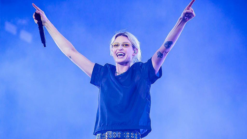 Emily Armstrong holding her arms up on stage. She is wearing a black t-shirt and black belt. She has blonde hair tied up, she is wearing yellow glasses.