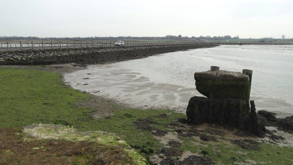 A single white car can be seen driving along a fenced causeway above tidal mudflats.