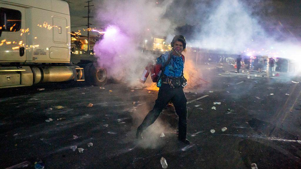 A lone demonstrator uses a fire extinguisher in the middle of the street, behind him a small barricade with people is visible. There's gas and smoke all over