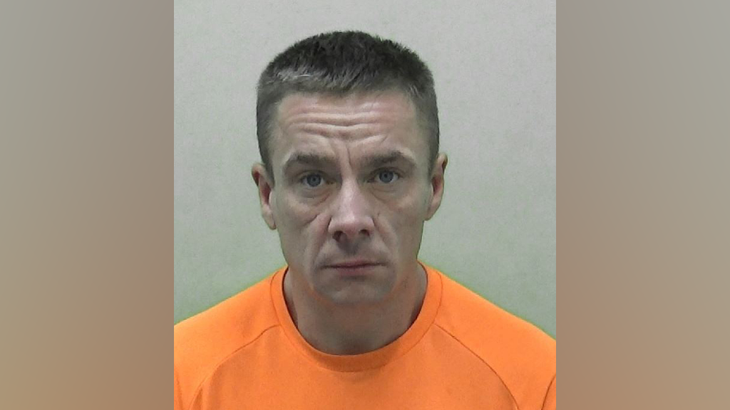 A mug shot of Christopher Donnison. He has short brown hair and is wearing an orange t-shirt. He is staring into the camera with a straight face.