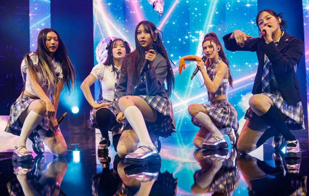 NewJeans on stage - the members are crouched down singing into their microphones. They are all wearing knee-length white socks, silver Mary Jane shoes and school uniform-style tartan skirts, white shirts and ties.