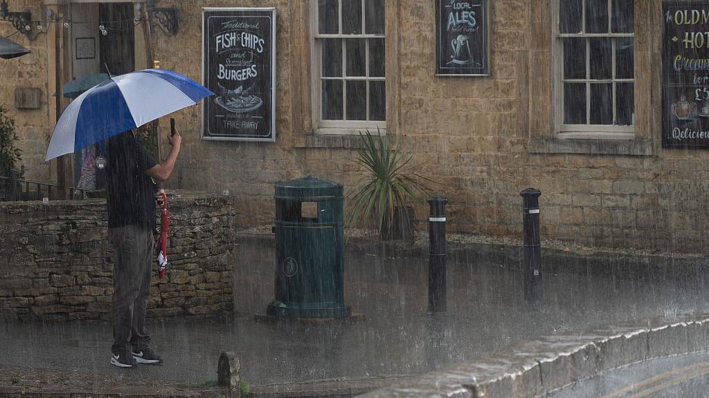photo of what appears to be a pub with a man holding an umbreall in one hand and his phone taking a picutre in the other.  There is pouring rain.