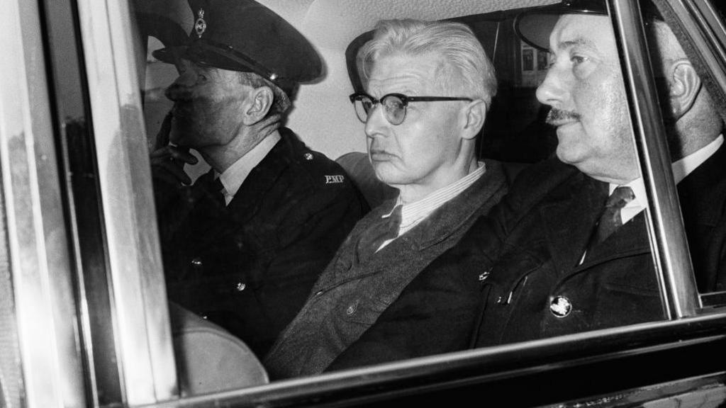 A black and white image showing a man with glasses and white hair sitting between two police officers in the back of a car in an image from the 1960s