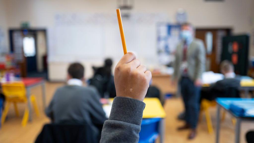 A close up of a student's raised hand with a classroom, students and a teacher in soft focus behind.