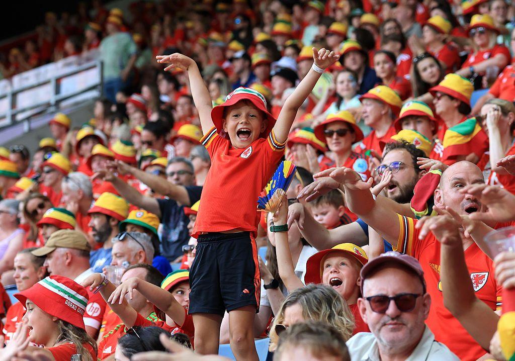 A boy stands over his seat at the stadium, cheering for Wales with his arms in the air. He is wearing a bucket hat and the Welsh jersey