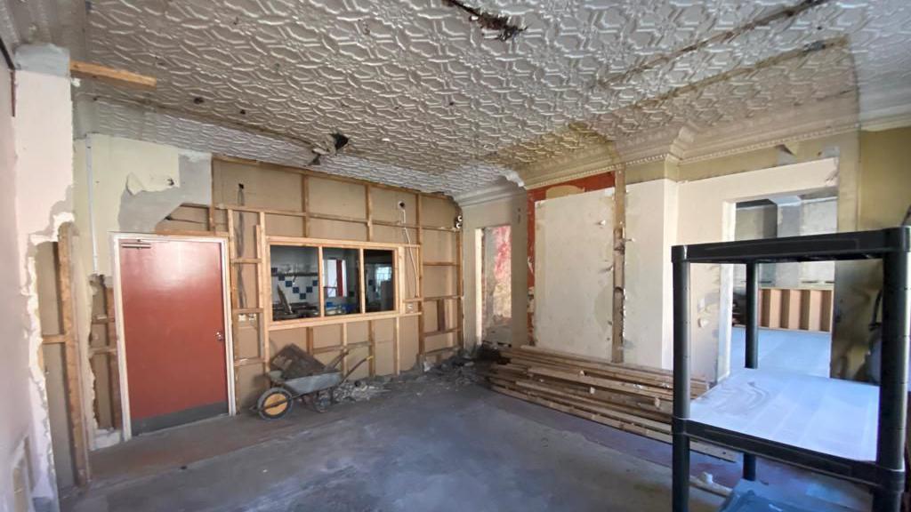 Interior of a room which has been stripped out. One wall appears to be completely plasterboard, a window area overlooks another room, and it has a red door. A wheelbarrow can be seen underneath the window. The two other visible walls have exposed plasterwork. The floor is bare and a metal stand  or shelving can bee seen on the right of the image.