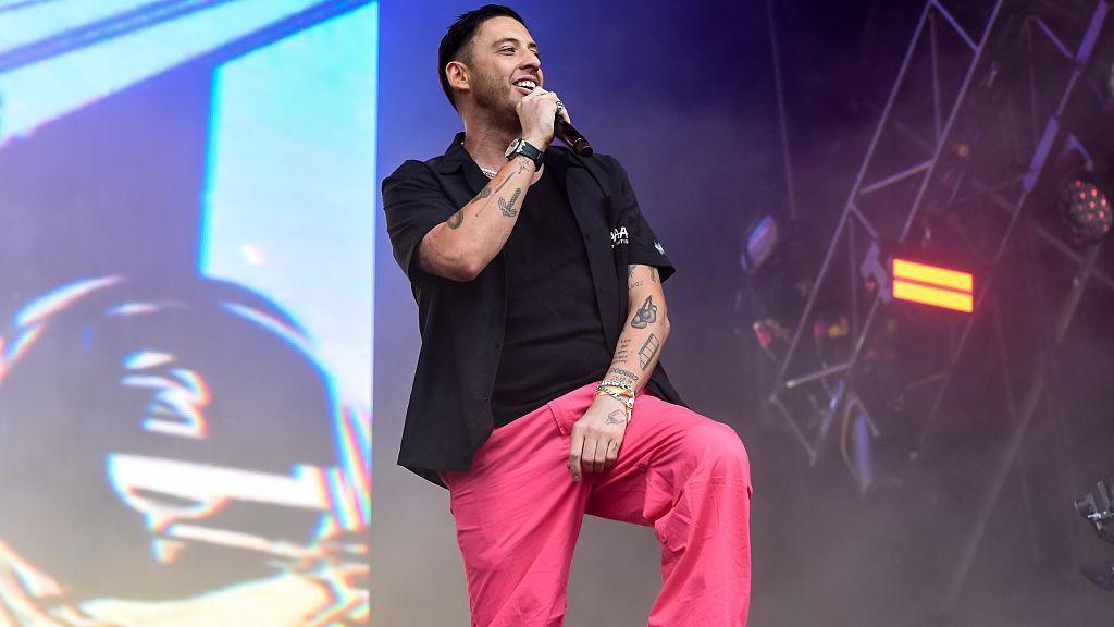 A man with dark hair and facial hair on stage singing into a handheld black microphone. He is wearing a black T-shirt, black shirt and pink trousers. He has tattoos on his arms.