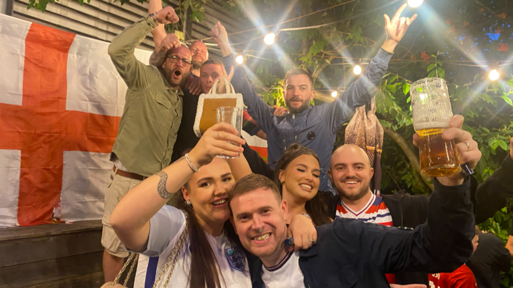 A group of England fans all raising their hands to cheer at the camera, they are in a pub garden and a St George's flag hangs behind them