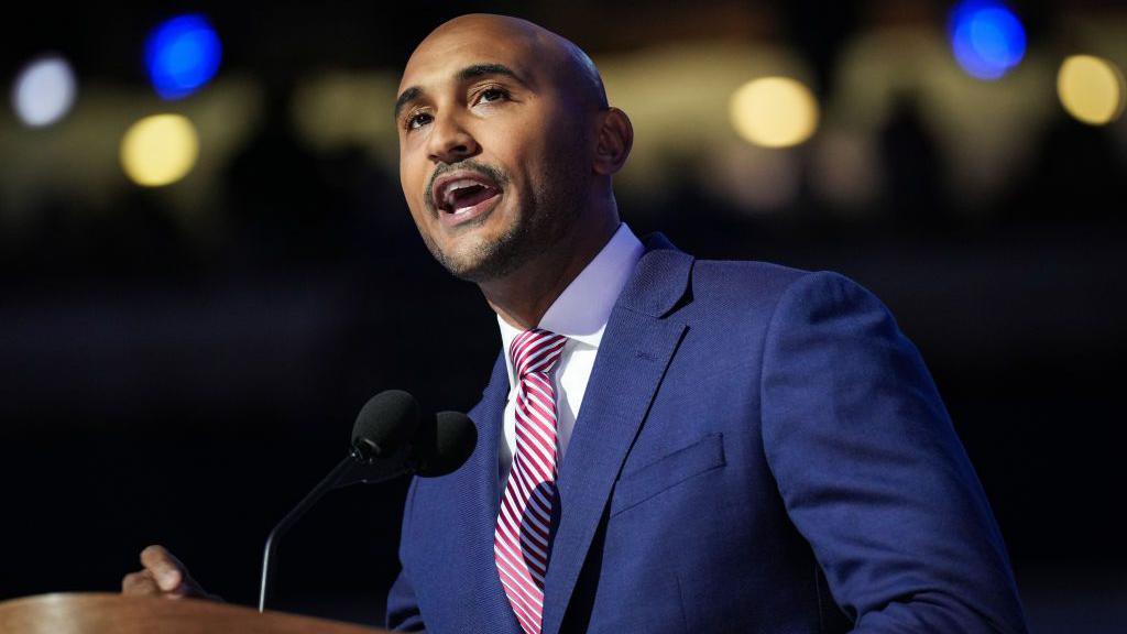 Shomari Figures, a bald black man with slight facial stubble, wearing a blue suit, white shirt and striped tie, speaks at the podium during the Democratic National Convention