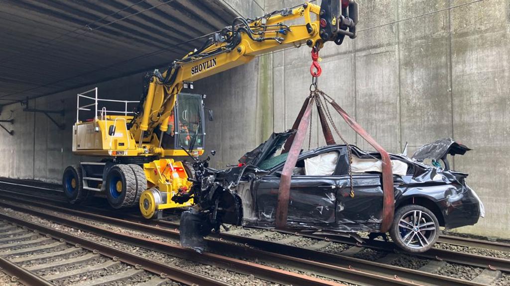 A yellow road-railer excavator with the crashed black BMW car slung from its crane arm